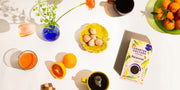 An overhead shot of 2 coffee mugs, a glass of juice, a blue glass case with 2 orange flowers and greenery, a green glass bowl of clementines, a yellow plate of pineberies, a sliced open blood orange, and checkered shortbread cookies surrounding a box of coffee named Perennial. Everything is on a cream colored background.